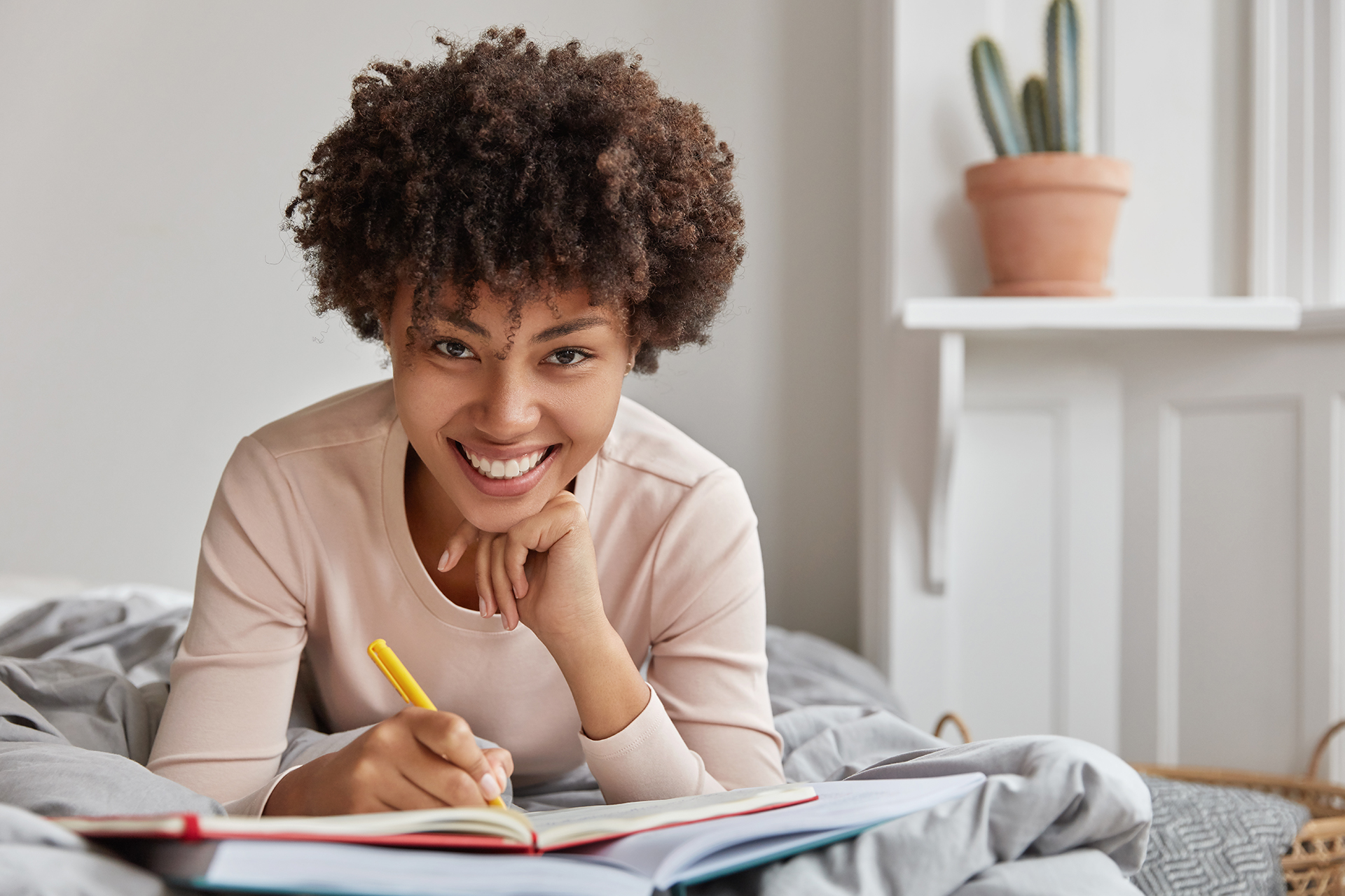 Photo of cheerful young Afro American lady writes down information in notebook with pen, records plans and notes checklist, lies at bed, holds hand under chin, spends weekend at home in bedroom